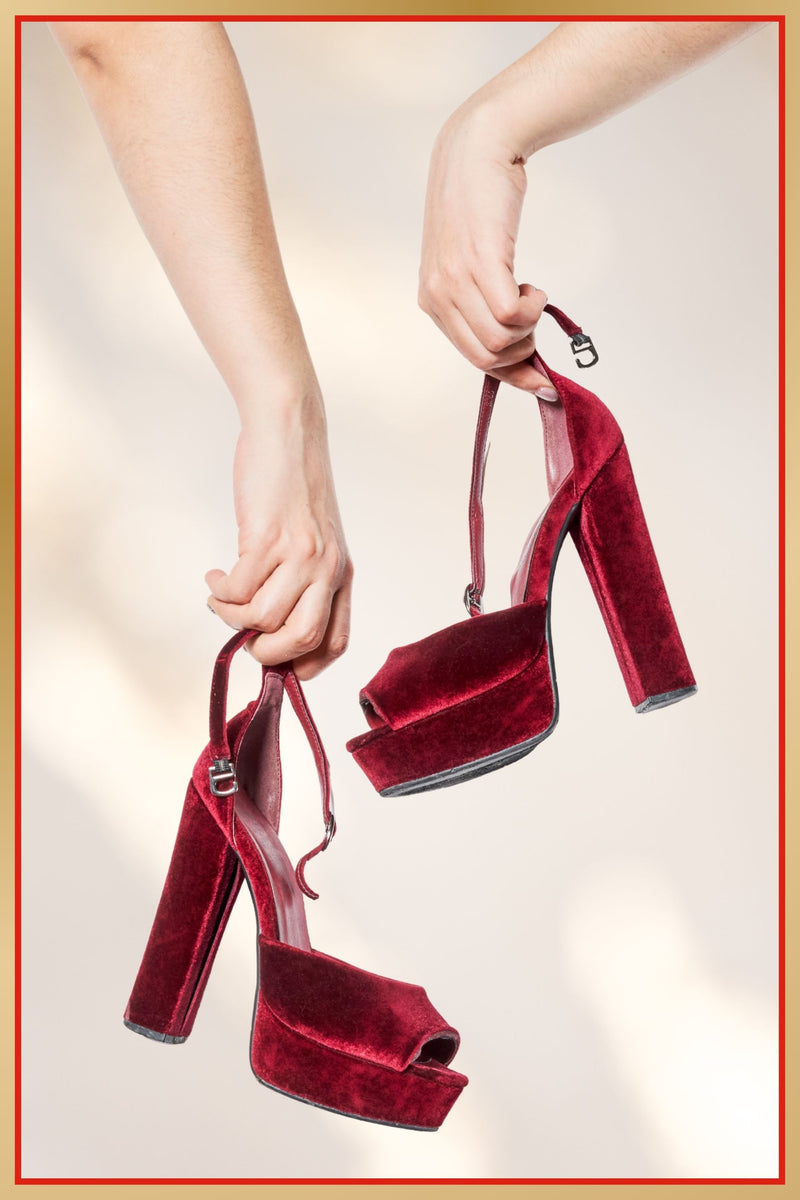 Red velvet high-heeled shoes held by a hand against a neutral background